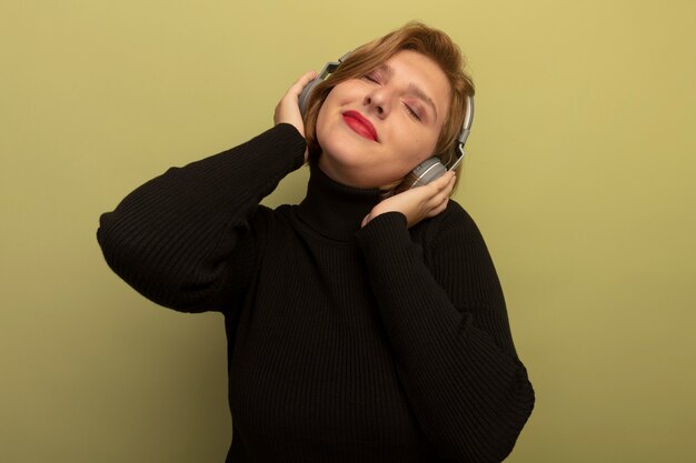 Heureuse jeune femme blonde portant et mettant les mains sur des écouteurs écoutant de la musique les yeux fermés isolés sur un mur vert olive avec espace de copie