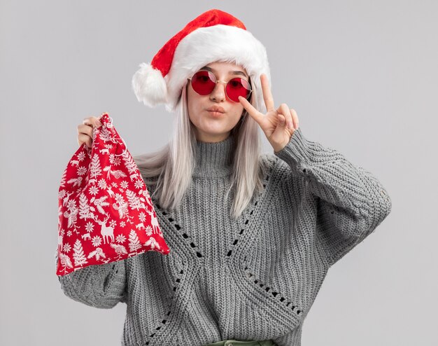 Heureuse jeune femme blonde en pull d'hiver et bonnet de noel tenant un sac rouge santa avec des cadeaux de noel montrant v-signe debout sur un mur blanc