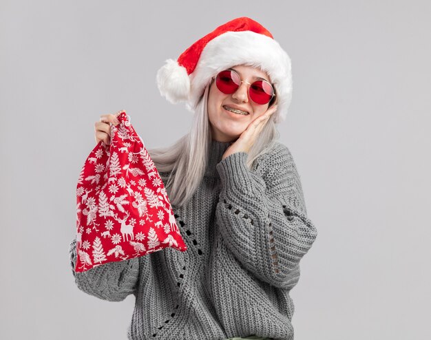 Heureuse jeune femme blonde en pull d'hiver et bonnet de noel tenant un sac rouge santa avec des cadeaux de noel souriant joyeusement debout sur un mur blanc