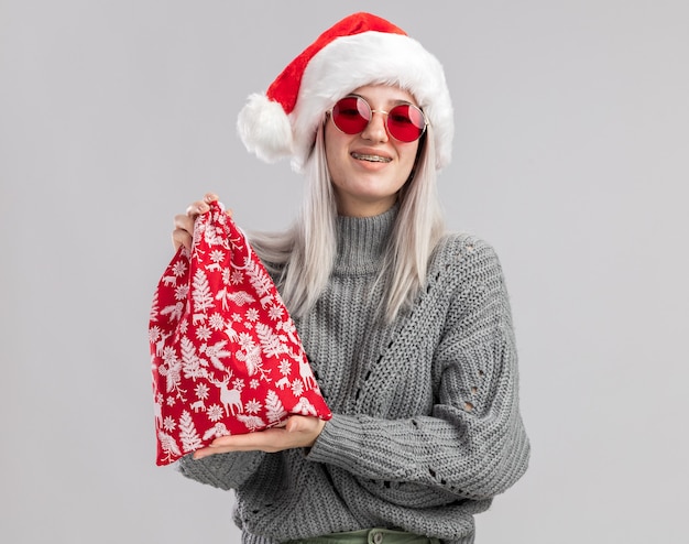 Heureuse jeune femme blonde en pull d'hiver et bonnet de noel tenant un sac rouge santa avec des cadeaux de noel souriant joyeusement debout sur un mur blanc