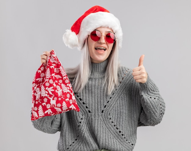 Heureuse jeune femme blonde en pull d'hiver et bonnet de noel tenant un sac rouge santa avec des cadeaux de noel souriant joyeusement montrant les pouces vers le haut debout sur un mur blanc