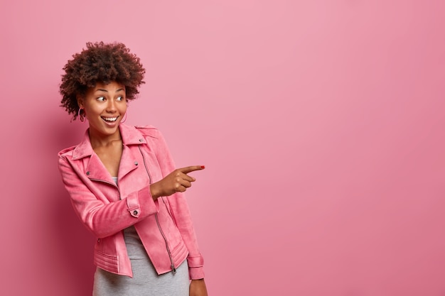 Heureuse jeune femme bouclée à la peau sombre indique à distance voit quelque chose d'étonnant rire positivement