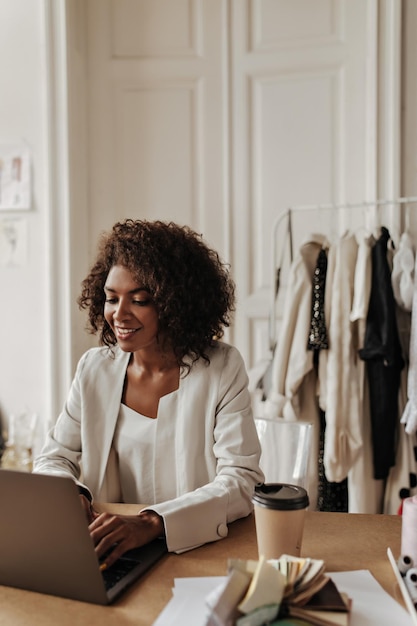 Heureuse Jeune Femme Bouclée En Veste Beige Haut élégant Semble Joyeux Assis Près Du Bureau Avec Une Tasse De Café Dessus Et Travaille Dans Un Ordinateur Portable Au Bureau