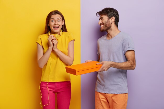 Heureuse jeune femme brune joyeuse garde les mains ensemble, porte un t-shirt décontracté jaune et un pantalon rose, reçoit une boîte en carton de petit ami