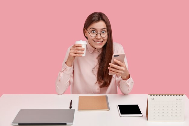 Heureuse jeune femme brune tient un téléphone portable moderne, types des messages texte, porte du café à emporter à la main, utilise le bloc-notes pour enregistrer des informations