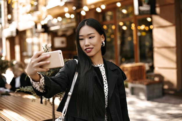 Heureuse jeune femme brune en trench-coat noir élégant sourit sincèrement, tient le téléphone et prend un selfie à l'extérieur près du café de la rue