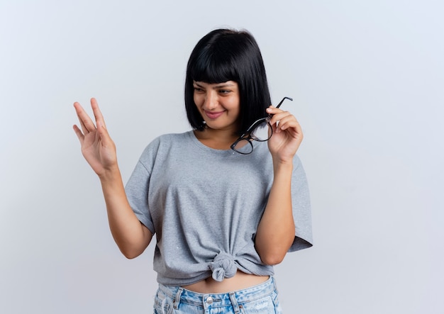 Heureuse jeune femme caucasienne brune tient des lunettes optiques et lève la main en regardant de côté