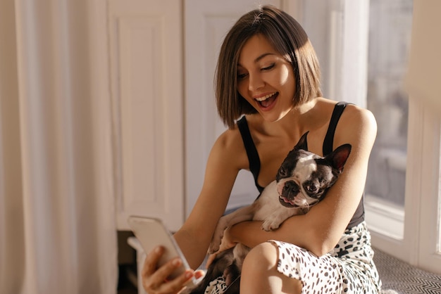Heureuse jeune femme caucasienne tenant un bouledogue français et faisant un appel vidéo via un smartphone à l'intérieur Concept d'amitié entre l'homme et les animaux