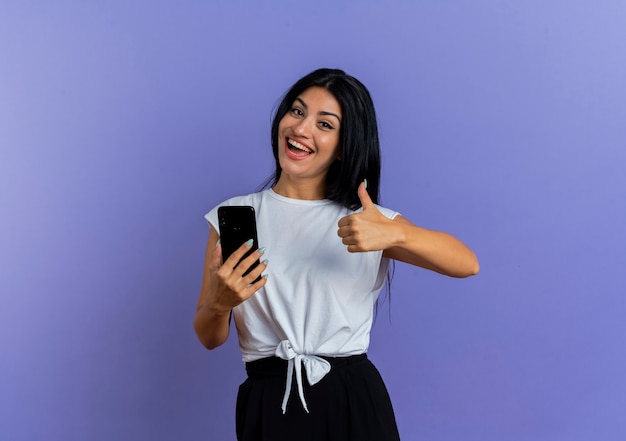 Heureuse jeune femme caucasienne tient le téléphone et les pouces vers le haut