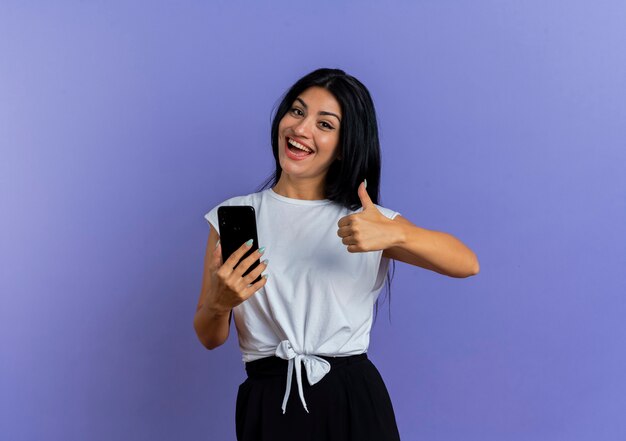 Heureuse jeune femme caucasienne tient le téléphone et les pouces vers le haut