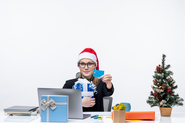 Heureuse jeune femme avec chapeau de père Noël et portant des lunettes assis à une table tenant un cadeau de Noël et montrant une carte bancaire sur fond blanc