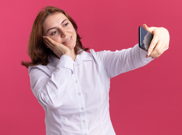 Heureuse jeune femme en chemise blanche à l'aide de smartphone faisant selfie souriant joyeusement debout sur le mur rose