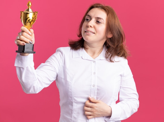 Heureuse Jeune Femme En Chemise Blanche Tenant Le Trophée D'or En Le Regardant Avec Une Expression Confiante Debout Sur Un Mur Rose