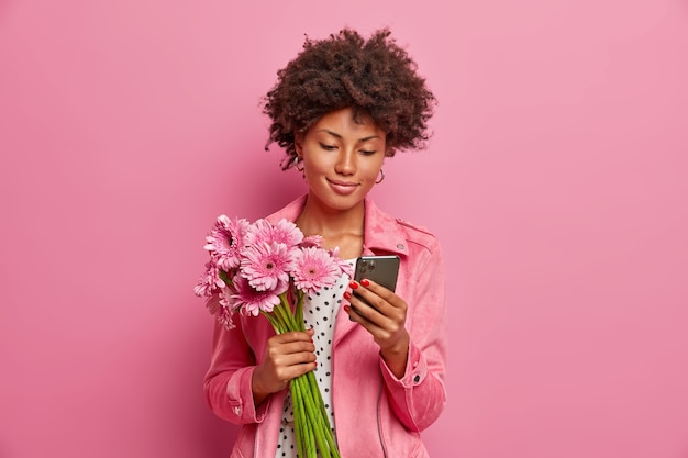 Photo gratuite heureuse jeune femme a les cheveux afro, a un bouquet de gerbera comme cadeau, pose avec de belles fleurs et un smartphone dans les mains, envoie des messages en ligne, reçoit un cadeau surprise