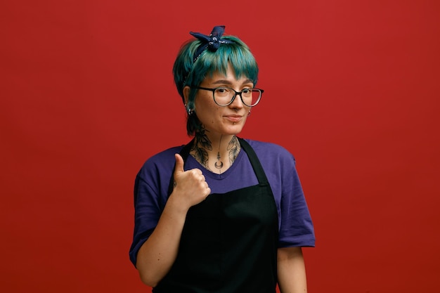 Photo gratuite heureuse jeune femme coiffeur portant des lunettes uniformes et un bandeau regardant la caméra montrant le pouce vers le haut isolé sur fond rouge