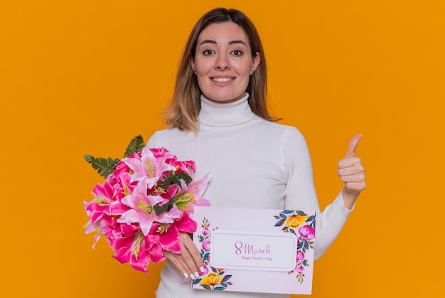 Heureuse jeune femme en col roulé blanc tenant une carte de voeux et un bouquet de fleurs