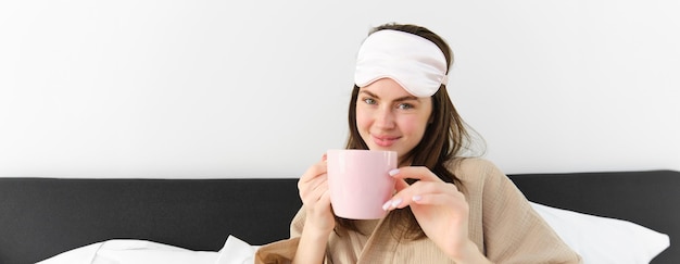 Photo gratuite heureuse jeune femme commençant sa matinée avec une tasse de café au lit en souriant et en profitant de sa journée de farniente