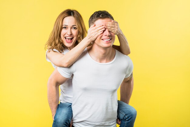 Heureuse jeune femme couvrant les yeux tout en chevauchant le dos de son petit ami sur fond jaune