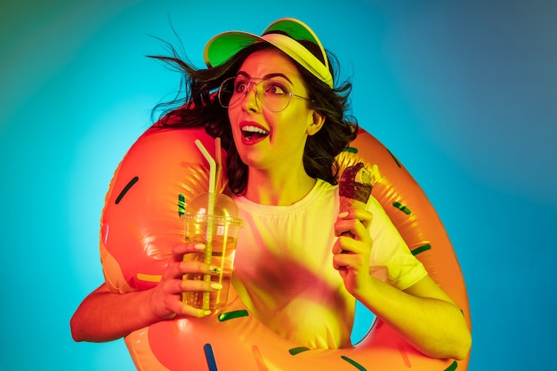 Heureuse jeune femme dans un anneau en caoutchouc avec une glace et une boisson sur néon bleu à la mode