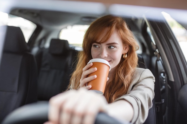 Heureuse jeune femme avec du café pour aller conduire sa voiture Femme sirotant un café en conduisant une voiture Jeune femme buvant du café en conduisant sa voiture Attrayant cheveux roux conduit une voiture