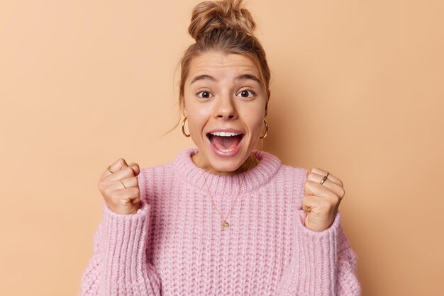 Heureuse jeune femme énergique serre les poings de joie s'exclame et se sent comme le gagnant célèbre la réussite a les cheveux peignés porte un pull tricoté isolé sur fond beige racines pour quelqu'un