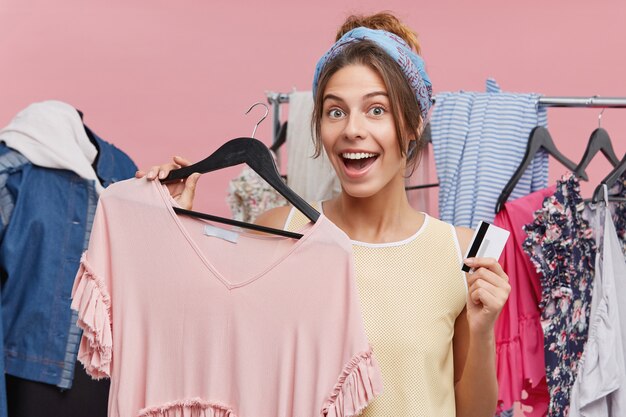 Heureuse jeune femme européenne accro du shopping se sentant excité lors de ses achats au centre commercial de la ville et avoir la chance de se rendre à la vente finale, tenant un cintre avec top tendance et carte de crédit, sur le point de l'acheter