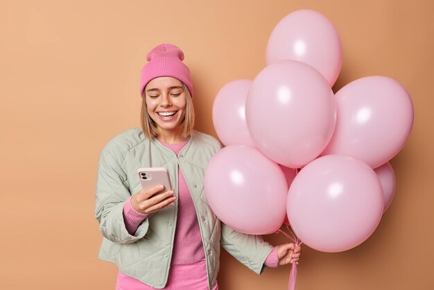 Heureuse jeune femme européenne souriante a une bonne humeur festive hods tas de ballons gonflés utilise des conversations téléphoniques avec des amis porte un chapeau et une veste vient à la fête isolée sur fond marron