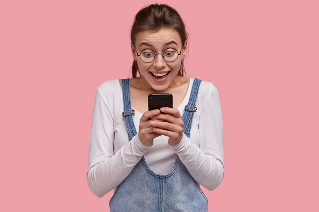 Heureuse jeune femme européenne tient un téléphone portable moderne, regarde une superbe invitation à faire la fête reçue via Internet, lit une bonne nouvelle impressionnante