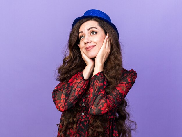 Heureuse jeune femme de fête portant un chapeau de fête gardant les mains sur le visage en regardant le côté isolé sur le mur violet
