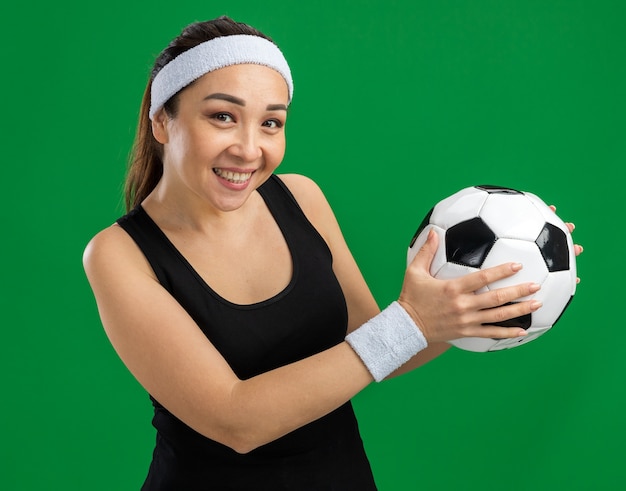 Heureuse jeune femme fitness avec bandeau tenant un ballon de football souriant joyeusement