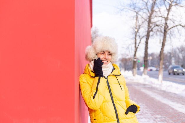 Heureuse jeune femme sur fond d'un mur rouge dans des vêtements chauds sur une journée ensoleillée d'hiver souriant et parlant au téléphone sur un trottoir de la ville enneigée