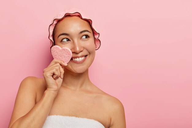 Heureuse jeune femme fraîche nettoie le visage avec une éponge, a une expression heureuse rêveuse, porte un bonnet de bain, se souvient d'un moment agréable en mémoire