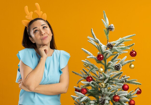 Heureuse jeune femme en haut bleu portant une jante drôle avec des cornes de cerf jusqu'à la pensée positive debout à côté d'un arbre de Noël sur fond orange