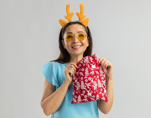 Heureuse jeune femme en haut bleu portant une jante drôle avec des cornes de cerf et des lunettes jaunes tenant un cadeau de Noël à la recherche d'un grand sourire sur le visage