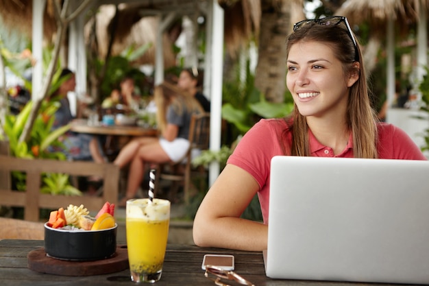 Heureuse jeune femme indépendante bénéficiant d'une connexion Internet sans fil gratuite assis devant un ordinateur portable générique au café en plein air. Femme joyeuse à l'aide d'un ordinateur portable pendant le déjeuner au restaurant avec terrasse