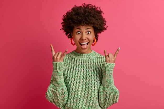 Photo gratuite heureuse jeune femme insouciante et rebelle à la peau foncée aime la musique géniale, fait un geste rock n roll, s'amuse au festival de musique ou à un événement cool, porte un pull décontracté, pose contre le mur rose.