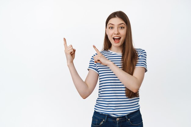 Heureuse jeune femme invitant et montrant le produit, pointant du doigt dans le coin supérieur gauche, souriant étonné sur blanc