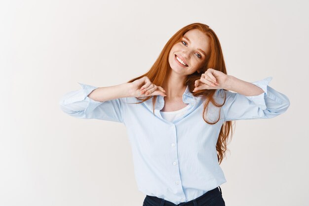 Heureuse jeune femme avec un long hiar rouge et des yeux bleus étirant les mains souriant joyeusement à la caméra debout sur fond blanc