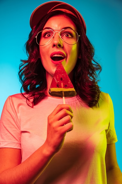 Heureuse jeune femme à lunettes de soleil manger des bonbons et souriant sur néon bleu à la mode