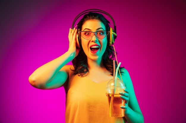 Heureuse jeune femme en lunettes de soleil rouges boire et écouter de la musique sur néon rose à la mode