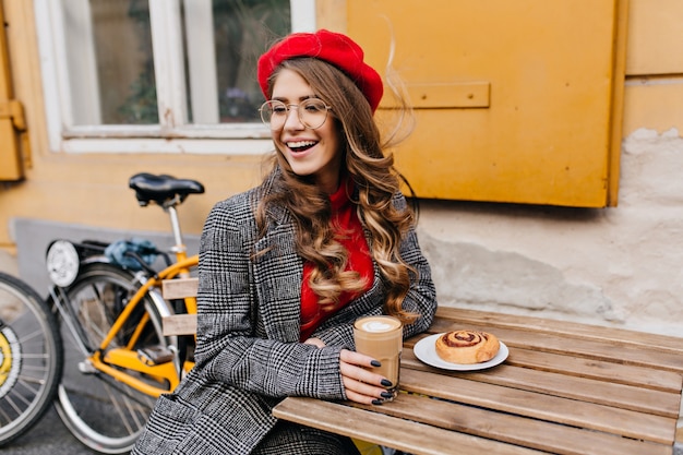 Heureuse jeune femme en manteau de tweed assis dans un café en plein air avec verre de cappuccino