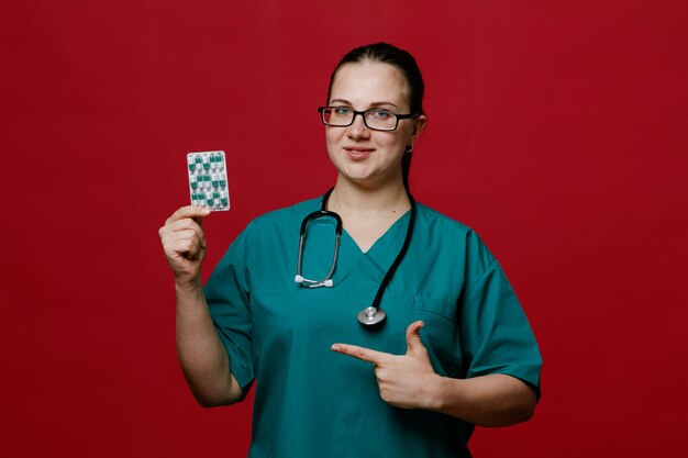 Heureuse jeune femme médecin portant des lunettes uniformes et un stéthoscope autour du cou regardant la caméra montrant un paquet de capsules pointant dessus isolé sur fond rouge