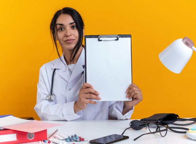 Heureuse jeune femme médecin portant une robe médicale avec stéthoscope est assise à table avec des outils médicaux tenant un presse-papiers isolé sur un mur jaune
