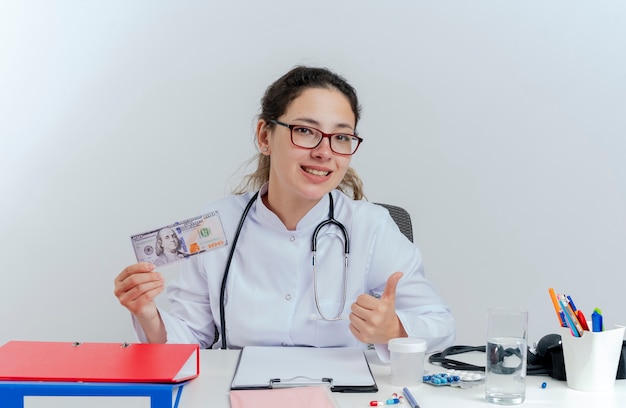 Heureuse jeune femme médecin portant une robe médicale et un stéthoscope et des lunettes assis au bureau avec des outils médicaux tenant de l'argent à la recherche de pouce vers le haut isolé