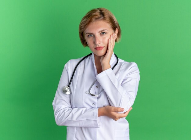 Heureuse jeune femme médecin portant une robe médicale avec stéthoscope mettant la main sur la joue isolée sur un mur vert