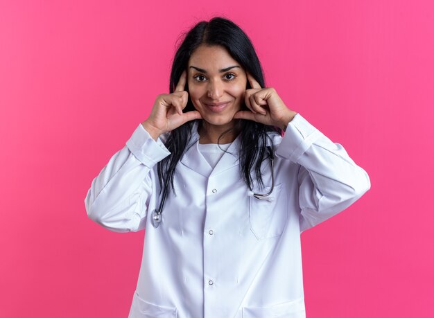 Heureuse jeune femme médecin portant une robe médicale avec stéthoscope mettant les mains sur le visage isolé sur un mur rose