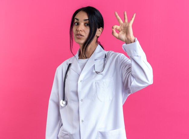 Heureuse jeune femme médecin portant une robe médicale avec stéthoscope montrant un geste correct isolé sur un mur rose