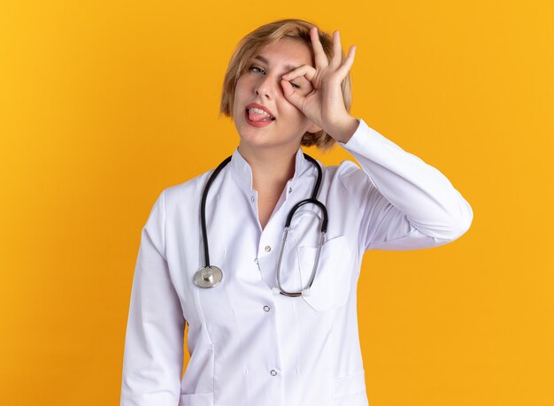 Heureuse jeune femme médecin portant une robe médicale avec stéthoscope montrant un geste de regard isolé sur un mur orange