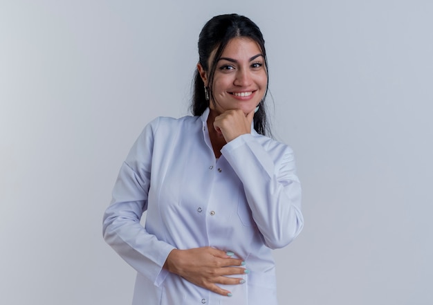 Heureuse jeune femme médecin portant une robe médicale à toucher le menton et le ventre isolé