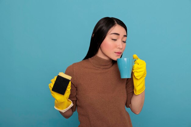 Heureuse jeune femme de ménage asiatique heureuse portant des gants jaunes prenant du café tenant une éponge Concept de maison de nettoyage, Thé de rafraîchissement à l'heure de la pause, Femme se sentant joyeuse satisfaite et positive